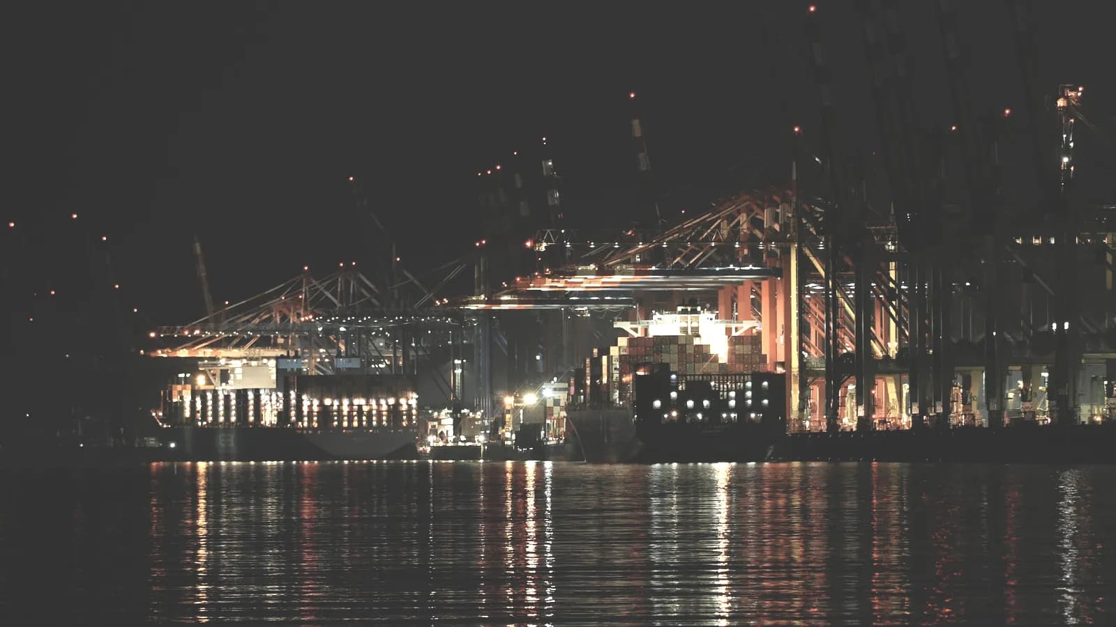 Terminal à conteneurs éclairé la nuit : portiques de quai en activité, piles de conteneurs et porte-conteneurs en cours de chargement.