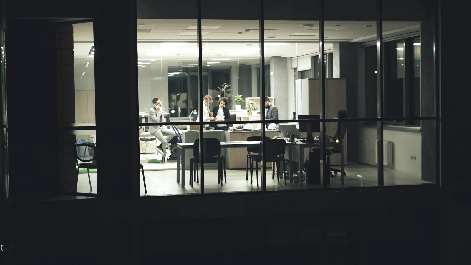 Open space éclairé la nuit, vu de l’extérieur à travers une baie vitrée sombre : quelques personnes travaillent encore autour d’un bureau, le reste du plateau est dans la pénombre.